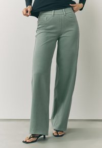 Nohavice s širokými nohavicami z svetlozelenej látky, s prednými vreckami, pútkami na opasok a jemnými švami. Kombinované s čiernymi sandálmi na podpätku.