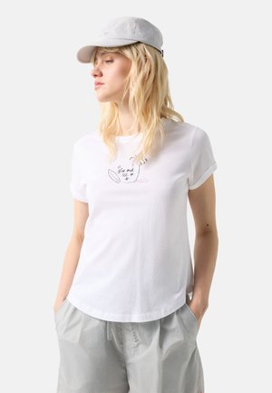 Blondynka w białej czapce, białej koszulce z napisem "Fire and Ice" oraz szkicami, i jasnoszarych luźnych spodniach, stojąca z rękami w kieszeniach.