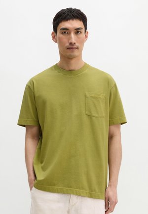 Mężczyzna ubrany w oliwkowo zielony T-shirt z krótkim rękawem i kieszenią na piersi oraz jasne spodnie, stojący na jednolitym białym tle.