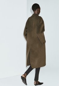 Cappotto in lana marrone con vestibilità relaxed, fessure laterali e colletto a punte, abbinato a scarpe nere slip-on e pantaloni skinny neri.