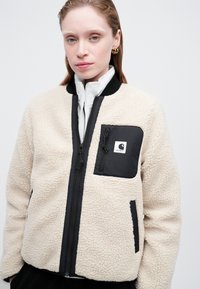 Kermanvärinen fleece-takki mustilla yksityiskohdilla, jossa on vetoketjulla varustettu etuosa, kaksi sivutaskua ja rintatasku logotiedolla.