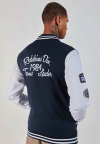 Veste de l'école navire et blanche avec des poignets rayés, présentant un texte brodé "Redskins Div. 1984 Team Leader" et des écussons sur les manches.