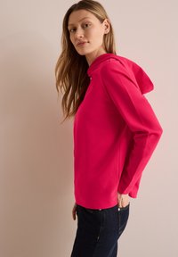 Sudadera rosa neón con una textura suave, estilo ajustado y un pequeño detalle de logo en la capucha. Combinada con unos pantalones vaqueros azul oscuro.