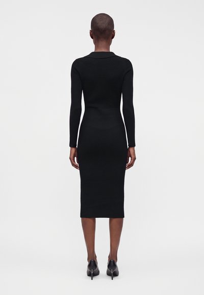 Femme aux cheveux courts portant une robe midi noire ajustée à manches longues et des chaussures à talons hauts noires, debout de dos devant un fond uni.