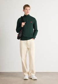 Dunkelgrüner Strickpullover mit einem Viertelreißverschluss, kombiniert mit beigefarbenen weiten Hosen. Schwarze Umhängetasche mit roten Akzenten, weiße Sneakers.