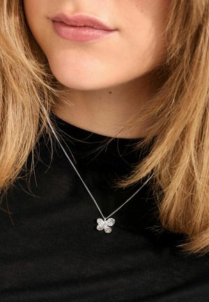 Collier en argent avec un pendentif en forme de fleur à quatre pétales, présentant un design texturé et des strass, porté sur un haut noir.