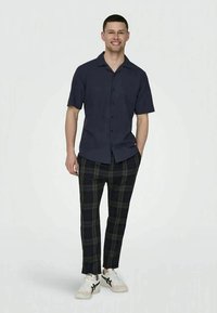 Homem a sorrir, vestindo uma camisa de manga curta azul-marinho, calças xadrez pretas e cinzas, e ténis brancos com riscas pretas, de pé com as mãos nos bolsos.