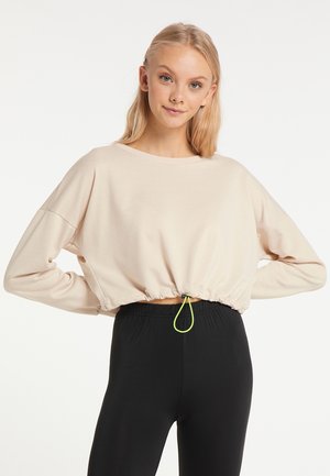 Blondynka z długimi włosami, ubrana w beżową krótka bluzę z neonowym sznurkiem i czarne legginsy z wysokim stanem, stoi z rękami na biodrach.