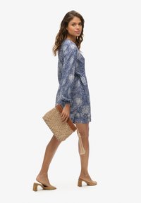 Vestido navy con manchas blancas, de mangas largas y dobladillo rizado. Sosteniendo un bolso de mano texturizado en tono beige con un borlón. Sandalias de tacón cuadrado en color beige.
