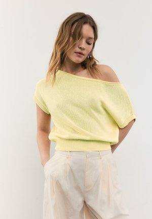 Ung kvinde med bølget hår iført en lysegul skulderfri kortærmet sweater og cremefarvede højtaljede bukser, hænderne i lommerne.