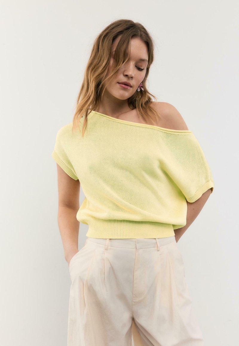 Jeune femme aux cheveux ondulés portant un pull jaune clair à manches courtes et épaules dénudées, et un pantalon crème taille haute, mains dans les poches.
