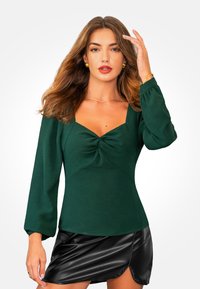 Top de manga larga, verde profundo, con escote cuadrado y detalle de torcido en el centro. Llevado con una mini falda negra y brillante.