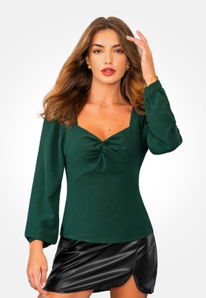 Top de manga larga, verde profundo, con escote cuadrado y detalle de torcido en el centro. Llevado con una mini falda negra y brillante.