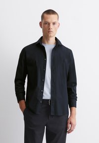 Zwarte button-up shirt met een spread collar, lange mouwen en een gladde textuur, gedragen over een lichtblauwe crewneck t-shirt.