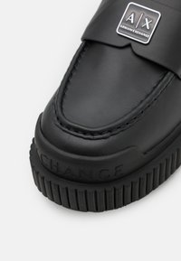 Scarpa slip-on in pelle nera con tomaia cucita, suola in gomma texturizzata e logo argento Armani Exchange sulla fascetta.