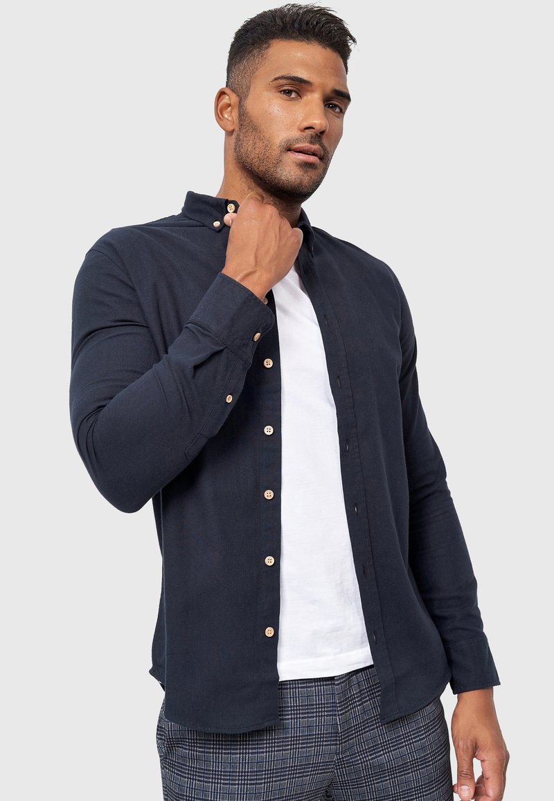 Marineblå skjorte med krave, prydet med træknapper og lange ærmer, iført over en hvid T-shirt, parret med mønstrede bukser.