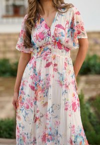 Femme portant une robe blanche à manches courtes avec des motifs floraux colorés rose, bleu et jaune, debout à l'extérieur.