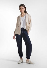 Beżowa lniana kurtka, biała koszulka z dekoltem w serek, granatowe spodnie dresowe z elastycznymi ściągaczami oraz kremowe trampki. Prosty i swobodny design.