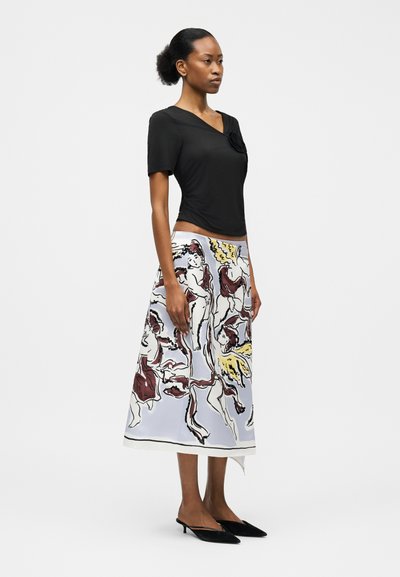 Γυναίκα που φοράει μαύρη μπλούζα με κοντά μανίκια, λευκή midi φούστα με πολύχρωμες απεικονίσεις αγγέλων και μαύρες γόβες με λουράκι πίσω, σε λευκό φόντο.