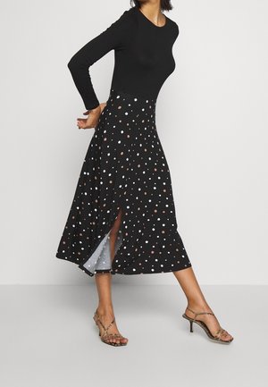 Frau trägt ein langärmeliges schwarzes Oberteil mit einem schwarzen, hoch taillierten Rock mit weißen und braunen Polka Dots und Schlitz, kombiniert mit Leopardenmuster-Pumps.