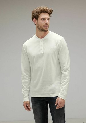 Pitkähihaisen valkoisen henley-paidan, jossa on kolmonen napitus, kevyesti textured ja huomaamaton logo rinnassa, yhdistettynä tummiin farkkuihin.