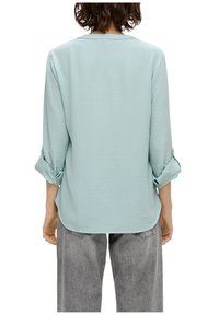 Personne portant un chemisier bleu clair à manches longues retroussées et un jean gris ample, vue de dos.