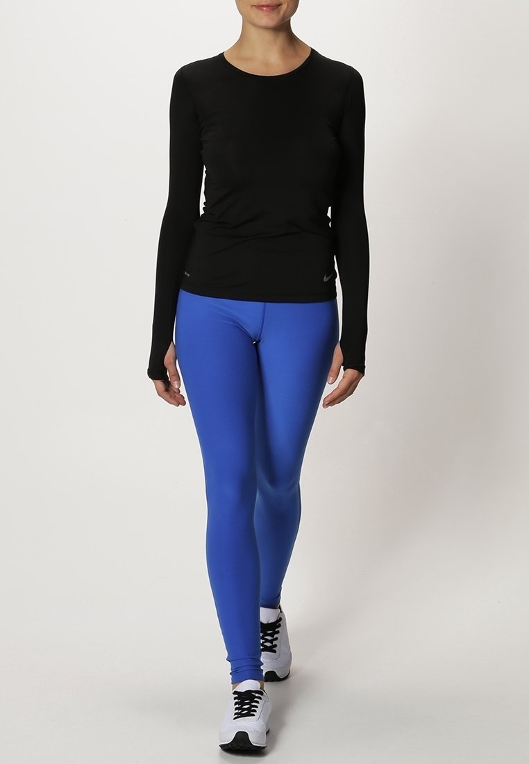 Černý dlouhý rukáv sportovní top v kombinaci s modrými legínami, vyrobený z elastického materiálu, s přiléhavým střihem a bílými teniskami.