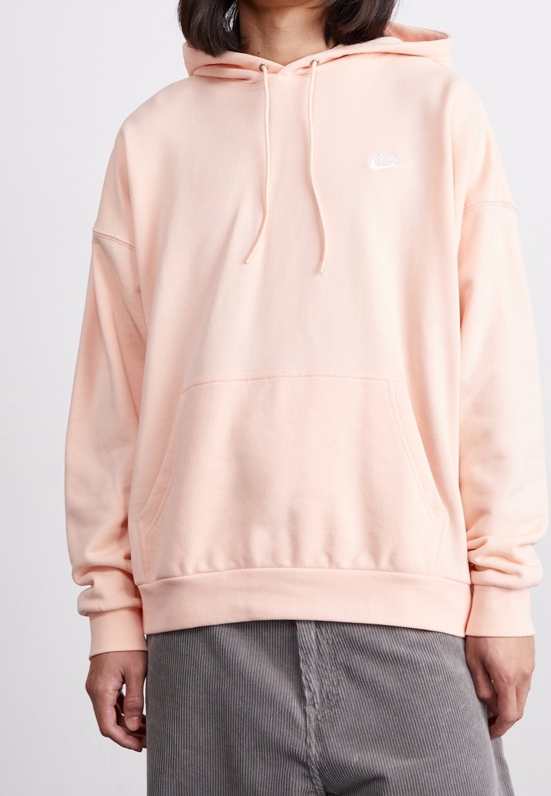 Ljusrosa hoodie med lös passform, dragsko, framficka och en liten vit Nike-logga på bröstet, tillverkad av mjukt bomullsmaterial.
