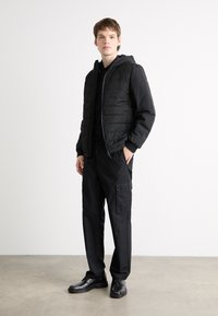 Giubbotto imbottito nero con cuciture testurizzate e cappuccio, indossato sopra un felpa nera. Abbinato a pantaloni cargo neri e scarpe nere.
