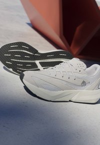 Chaussures de course blanches et grises avec semelles à crampons noires sur un sol clair près d'une sculpture rouge angulaire sous un soleil éclatant.