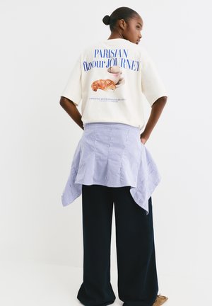 T-shirt graphique crème avec le texte "PARISIAN flavour JOURNEY" et une illustration de croissant, associé à une jupe portefeuille rayée bleue et un pantalon large sombre.
