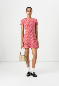 Robe rouge à fleurs avec des manches courtes, corsage ajusté et jupe évasée ; associée à un sac à main beige, des chaussettes blanches et des chaussures noires.