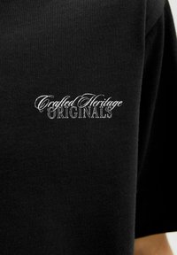 Czarna tkanina z białym haftowanym napisem "Crafted Heritage ORIGINALS" na obszarze klatki piersiowej odzieży.