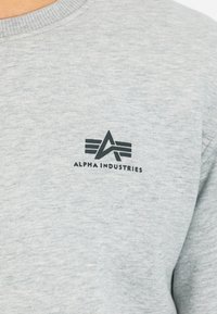 Hellgrauer Pullover aus weichem Stoff mit einem schwarzen Logo, das stilisierte Schriftzüge und ein grafisches Design auf der linken Brustseite zeigt.