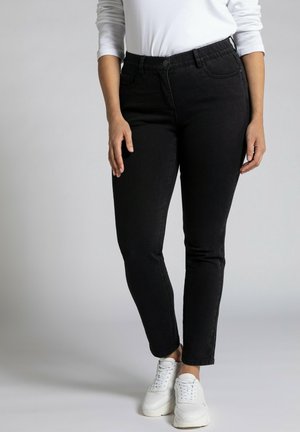 Personne portant un jean slim noir, une chemise blanche à manches longues et des baskets blanches, debout sur un fond clair uni.