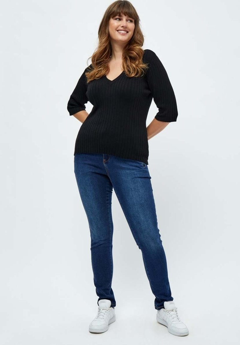 Schwarzes, geripptes Stricktop mit V-Ausschnitt und halblangen Ärmeln, kombiniert mit blauen, eng anliegenden Jeans und weißen Sneakers. Einfaches, lässiges Outfit.