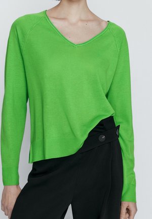 Maglione verde lavorato a maglia con scollo a V, maniche lunghe e polsini a coste. Indossato sopra pantaloni neri a vita alta con dettagli a bottoni.