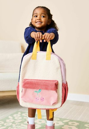 Mladá dívka v tmavě modrých šatech se usmívá a drží krémový a růžový batoh se žlutými rukojeťmi a motivem třešní.