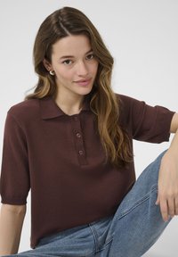 Polo shirt marrone a maniche corte con colletto, dotato di un'apertura con tre bottoni. Materiale in maglia morbida, abbinato a jeans in denim blu.