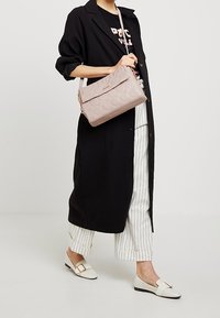 Quiltet lyserød skuldertaske med en frontklap, guldfarvet hardware og en lang rem, parret med en sort trenchcoat og stribede bukser.