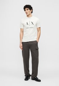 Άντρας που φοράει λευκό t-shirt με το λογότυπο Armani Exchange, σκούρο φαρδύ παντελόνι με πλαϊνή τσέπη και μαύρα παπούτσια, στέκεται με το ένα χέρι στην τσέπη.