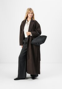 Kobieta w długim ciemno-brązowym płaszczu, białej bluzce, szerokich ciemnych spodniach, czarnych butach, trzymająca dużą czarną torbę, stojąca na tle białym.