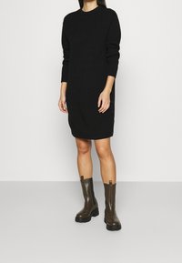 Donna che indossa un vestito a coste nero a maniche lunghe, lungo fino al ginocchio, con bottoni laterali e stivali alla caviglia spessi di colore marrone scuro, in piedi su uno sfondo neutro.