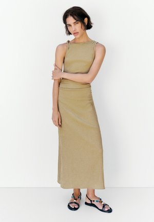 Donna con capelli corti e scuri che indossa un abito maxi beige senza maniche e sandali neri bassi con perline colorate, in piedi davanti a uno sfondo bianco.