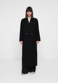 Cappotto lungo in misto lana nero con design doppiopetto, ampie rever e maniche lunghe. Presenta grandi bottoni e una silhouette dritta.