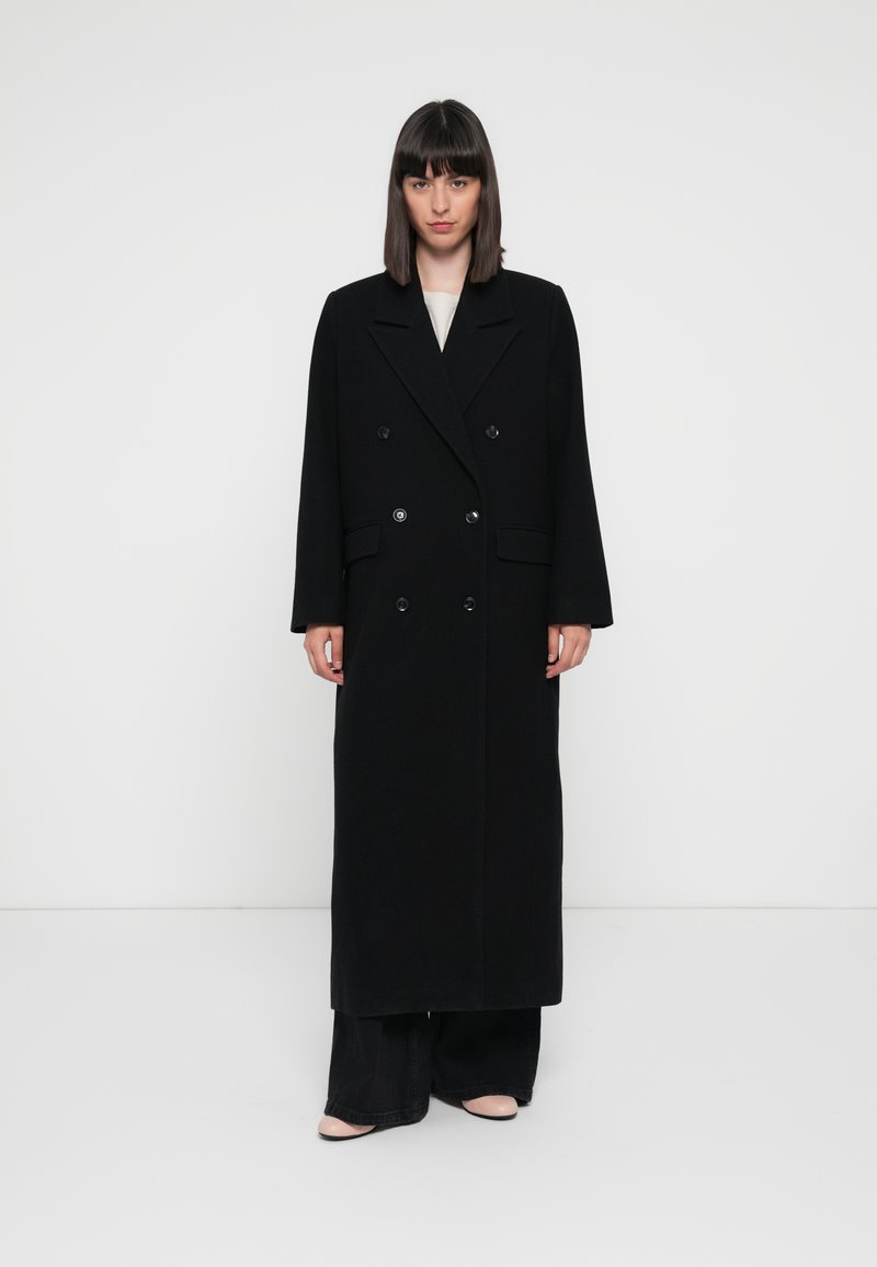 Cappotto lungo in misto lana nero con design doppiopetto, ampie rever e maniche lunghe. Presenta grandi bottoni e una silhouette dritta.