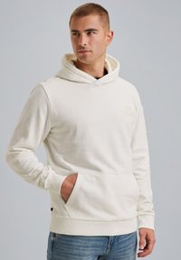 Krémová mikina vyrobená z mäkkej látky s klokanm vreckom, nastaviteľnou kapucňou so šnúrkou a nenápadným logom na hrudi.