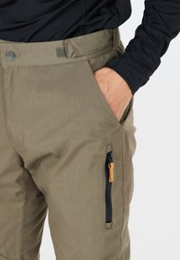 Pantalons olive vert résistants à l'eau en tissu durable, dotés d'une poche zippée avec une languette en cuir beige et de poches latérales.