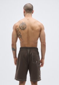 Hnedé plavkové šortky s bielym prešívaním, so zadným vreckom a elastickým pásom, zobrazené zozadu, s viditeľným tetovaním na pleci.