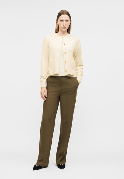 Γυναίκα που στέκεται με τα χέρια στις τσέπες, φορώντας κρεμ ζακέτα με χρυσά κουμπιά, ελαφρώς φαρδιά παντελόνια ελιάς και μαύρα παπούτσια με μυτερή μύτη πάνω σε λευκό φόντο.